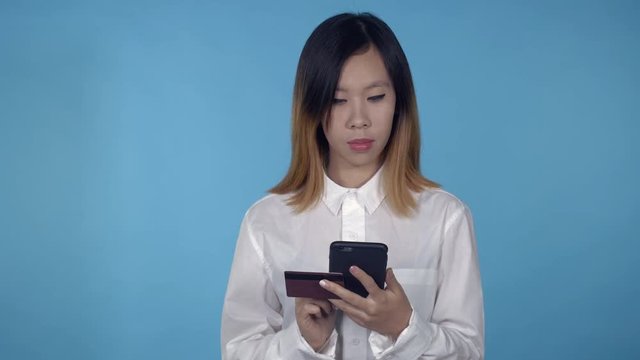 young asian woman using smartphone for buying online or pay order or bills on blue background in studio. attractive millennial girl wearing white casual shirt looking at the camera smiling