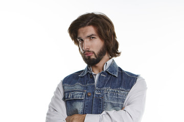 Dissatisfied young bearded man wearing denim vest over grey shirt keeping arms folded and staring at camera with suspicious distrustful look, posing at white studio wall with copy space for your text