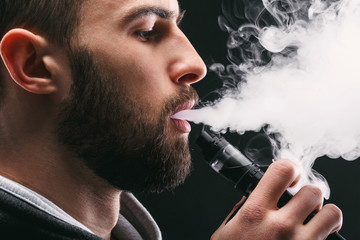 Young man vaping e-cigarette with smoke on black