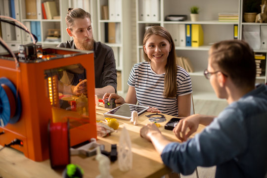 Portrait of three creative young people using 3D printer operating it via digital tablet studying for design project in college, focus on smiling woman in center