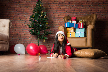 Little Indian girl writes letter to Santa or drawing on christmas day in santa clause attire and...