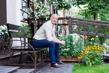 handsome man sit on the bench with garden background at spring time. green grass, jeans, yellow flowers, jeans, shirt, boots. summer