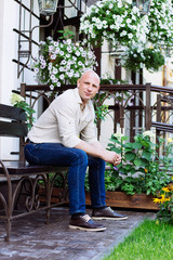 adult bald man sit on the bench at garden background at spring time. green grass, jeans, yellow flowers, jeans, shirt, boots. summer