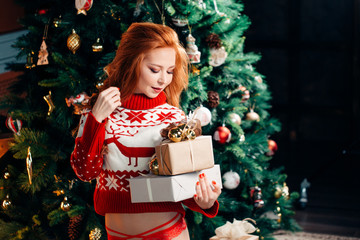 Sexy young woman in sweater and red panties hat is preparing for celebrating New Year at home. Holding gift box in hands on the background of beautiful Christmas tree.