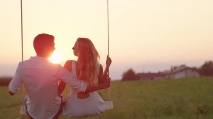 SLOW MOTION CLOSE UP: Joyous girlfriend and boyfriend kissing and laughing on swing on summer evening in countryside. Young couple enjoying last sunrays on romantic date whilst swaying on rope swing.