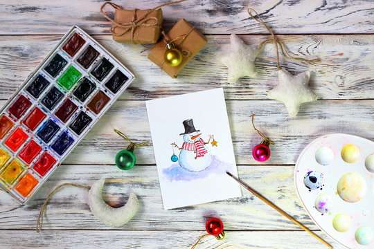 New Year (Christmas) cards with funny snowman on white boards background with watercolors and brush for drawing. Top view. Merry christmas and happy new year greeting card
