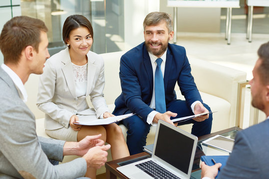 Group of successful business people working together in hall of modern office building focus on smiling young Asian woman and bearded mature man talking to young colleague sitting at coffee table