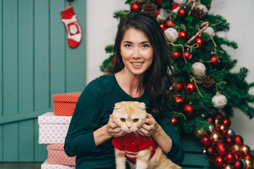 Portrait of young woman and cute cat wearing sweater. christmas tree and presents background