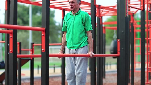 Elderly grey-haired sportsman doing physical exercise on sports ground. Active elderly athlete in green t-shirt trains in open air gym. Aged grey-haired man doing strenuous exercise on parallel bars