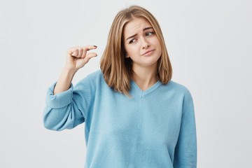 Fototapeta premium Pretty cute girl wearing casual clothes showing something small in size with hands while gesturing, frowning her brows. Blonde European female demonstrating size of something posing at camera