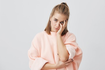 Bored European girl student wearing stylish pink sweatshirt touching face with hand, looking...