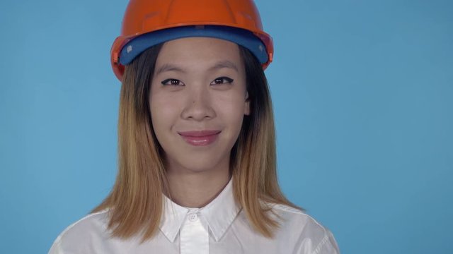 close up portrait young asian architect orange hard hat on blue background in studio. attractive millennial girl looking at the camera
