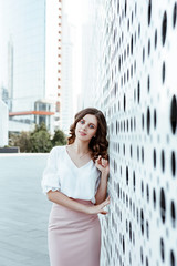 Office style. A business portrait of the young woman in a white blouse and a pink skirt against the background of city architecture.