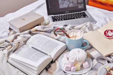 Fototapeta na wymiar Winter mood composition Christmas Day at home with cup of hot cacao or coffee with marshmallow sweets candy cane and lollipop read book films at laptop work in bed with wool knitted plaid New Year.