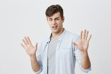 Fototapeta premium Offended caucasian young male has rush reaction on what he heard, raises hands, like saying stop that, has displeased and angry expression, isolated against gray background.