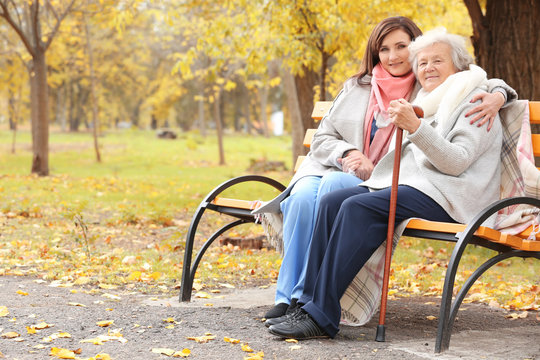 Senior woman with cane and young caregiver sitting on bench in park
