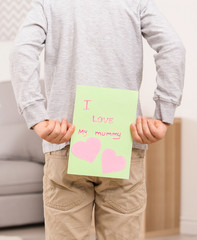 Cute little boy hiding greeting card for Mother's Day behind his back at home