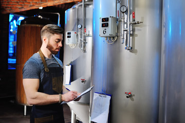 Fototapeta na wymiar A handsome bearded male brewer records data against a background of beer tanks. Brewing