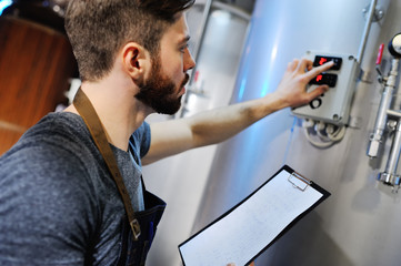 Fototapeta premium A handsome bearded male brewer records data against a background of beer tanks. Brewing