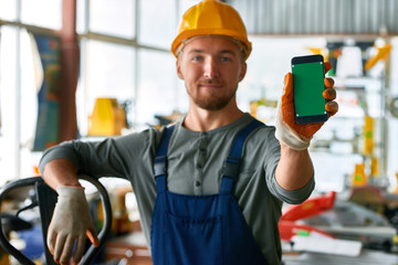 Portrait of cheerful young worker wearing hardhat posing looking at camera and holding smartphone...