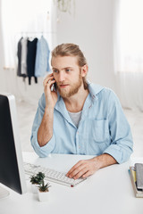 Vertical shot of caucasian handsome freelancer with beard and fair hair checking information and typing promotional text. Pleasant looking male employee has phone conversation with clients.