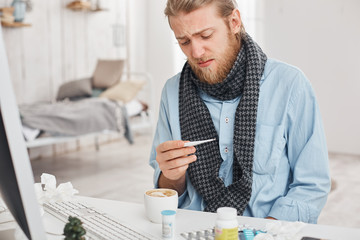 Ill or sick bearded male uses thermometer to measure temperature of his body. Fair-haired man desperately looks at thermometer, suffers from bad cold, surrounded by medicine at his workplace.