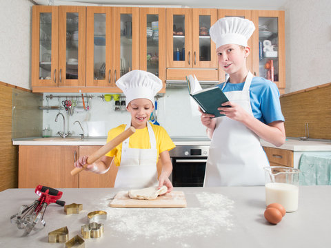 Little girl in the form of a chef with an apron and a cap blinks the dough with her hands, and next to her is her adult brother and reads a book on the background of the kitchen with a table