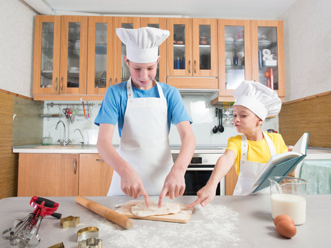 Schoolboy in the form of a cook in an apron and a cap holds a rolling pin in his hands and rolls out the dough, and next to it is his younger sister and reads a book on the background of the kitchen 
