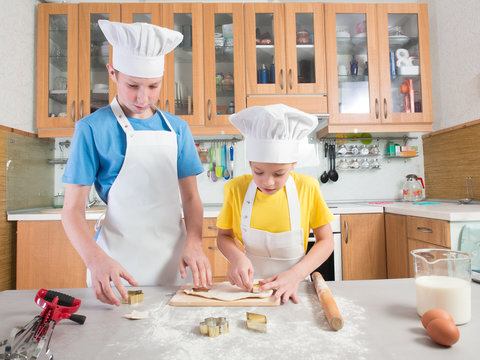 Schoolboy in the form of a cook in an apron and a cap together with his little sister they make pies from pastry on the background of the kitchen with a table 