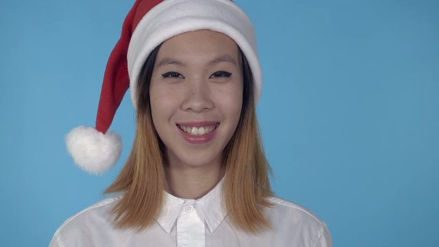close up portrait young asian woman posing wearing santa claus hat on blue background in studio. attractive millennial looking at the camera laughing
