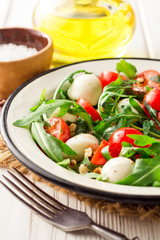Fresh salad with arugula, cherry tomatoes, mozzarella cheese and walnuts on white wooden background.