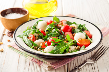 Fresh salad with arugula, cherry tomatoes, mozzarella cheese and walnuts on white wooden background.