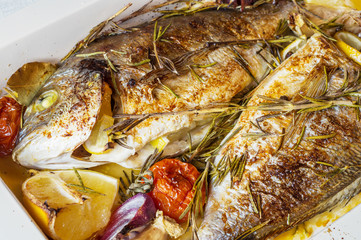 Two delicious whole baked fish. Baked dorado with lemon, onions, rosemary, cherry tomatoes, and spices on a wooden background. Delicious roasted dorado on oven tray. Diet and healthy food. Top view.