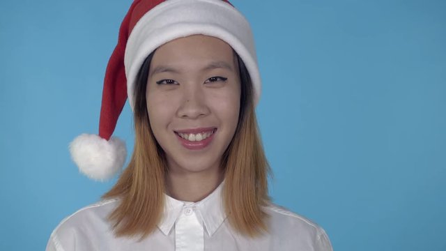 close up portrait young asian woman shows symbol like wearing santa claus hat on blue background in studio. attractive millennial looking at the camera