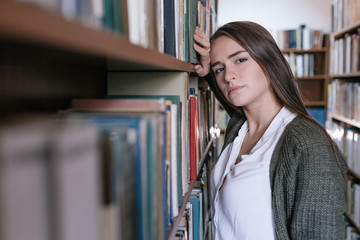 a beautiful young thin girl with a dark straight hair stands in the library and leans her head of books.