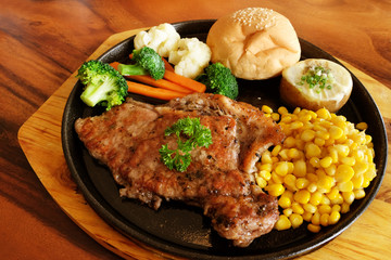 Meat steak with many side dish, bread with white sesame, corn, broccoli, cauliflower, carrot, black pepper and parsley leaves, served in hot black pan on wooden plate