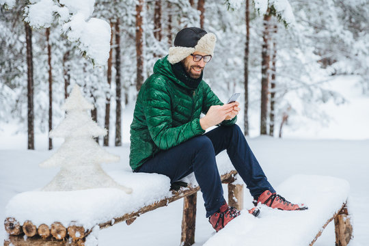 Cheerful male enjoys communication on smart phone and loneliness, calm atmosphere, sits on wooden bench covered with snow in winter forest, happy to recieve message from close friend