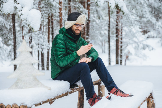 Smiling positive male wears warm winter clothes, reads message on mobile phone, spends free time in calm atmosphere outdoors in winter forest, enjoys fresh air. People, rest, technology concept