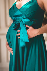 Closeup on tummy of pregnant woman, wearing long emerald color dress, holding in hands on belly. New life concept
