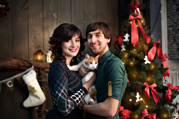 Obraz na płótnie Canvas Beautiful couple in love in green with a red cat on a background of Christmas tree decorations. Happy family in new year loft with red yellow bokeh. Wooden wall. Copy space. Close up portrait. 