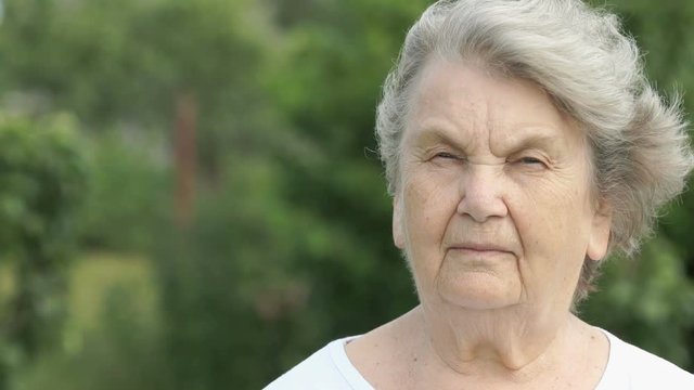 Portrait of serious mature elderly woman with gray hair dressed in a white t-shirt in the background of the green park in summer. Slow Motion