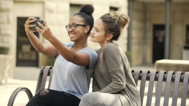 A pretty African American college girl and her Caucasian friend and colleague sitting outside on a campus bench taking a selfie.
