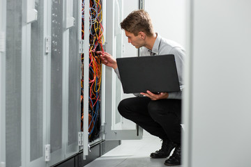 Portrait of modern young man holding laptop standing in server room working with supercomputer