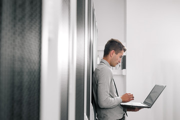 Portrait of modern young man holding laptop standing in server room working with supercomputer