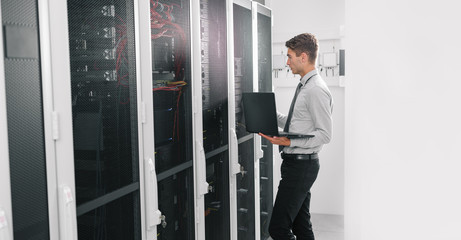 Portrait of modern young man holding laptop standing in server room working with supercomputer