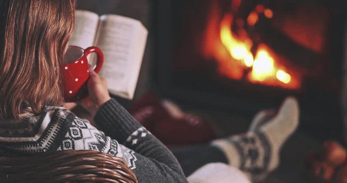 Woman Reading a Book by the Fireplace. SLOW MOTION 4K. Young woman reading a book by the warm fireplace decorated for Christmas. Relaxed holiday evening concept.