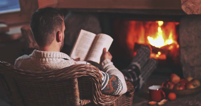 Man Reading a Book by the Fireplace. SLOW MOTION 4K. Young man reading a book by the warm fireplace decorated for Christmas. Relaxed holiday evening concept.