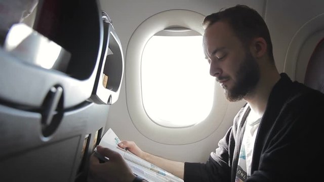 An unshaven man is reading a magazine in a plane near porthole. Male passenger learns the press during a flight in an air company.