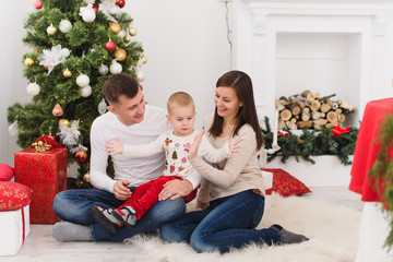 Happy young cheerful parents with cute little son. Child boy sitting in light room at home with decorated New Year tree and gift boxes. Christmas good mood. Family, love and holiday 2018 concept.