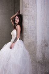 Fototapeta na wymiar Young beautiful woman in white wedding dress with bare shoulders posing near a concrete wall